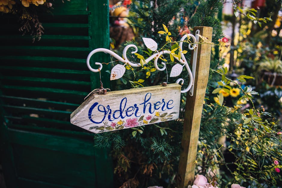 A rustic wooden sign with the word 'Order here' painted in blue, hanging from a decorative white metal bracket with scrollwork, amid lush green foliage and blooming flowers in shades of pink and yello