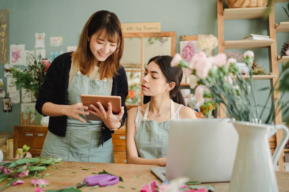 Photograph of two women inside a flower shop, with one standing and the other seated at a wooden worktable cluttered with fresh floral materials, including pink and purple petals and green stems. The
