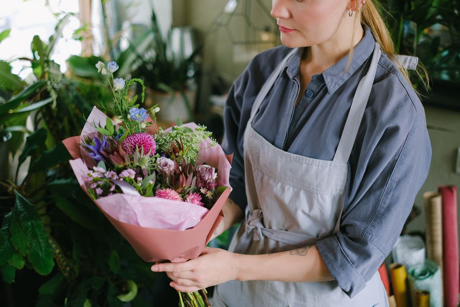 A woman in a blue shirt and gray apron holds a vibrant bouquet of fresh flowers in a florist's shop, featuring pink roses, purple lisianthus, white baby’s breath, and green foliage, wrapped in pink 