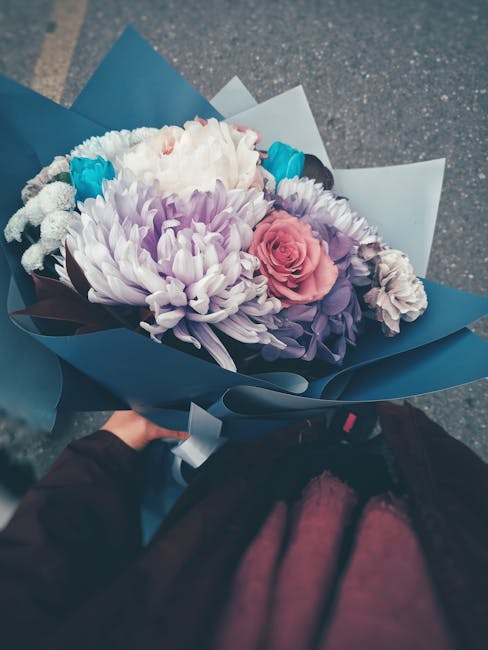 A bouquet of fresh flowers arranged with light lavender chrysanthemums, soft pink roses, white carnations, and accents of bright blue flowers, all wrapped in elegant blue and white paper and secured w