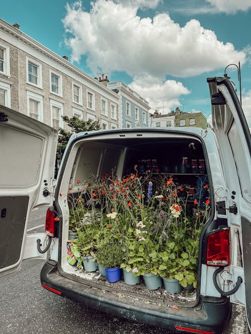 The open rear of a white delivery van filled with potted flowers and plants, including red, white, and green foliage, situated on a city street with classic white terraced buildings under a partly clo