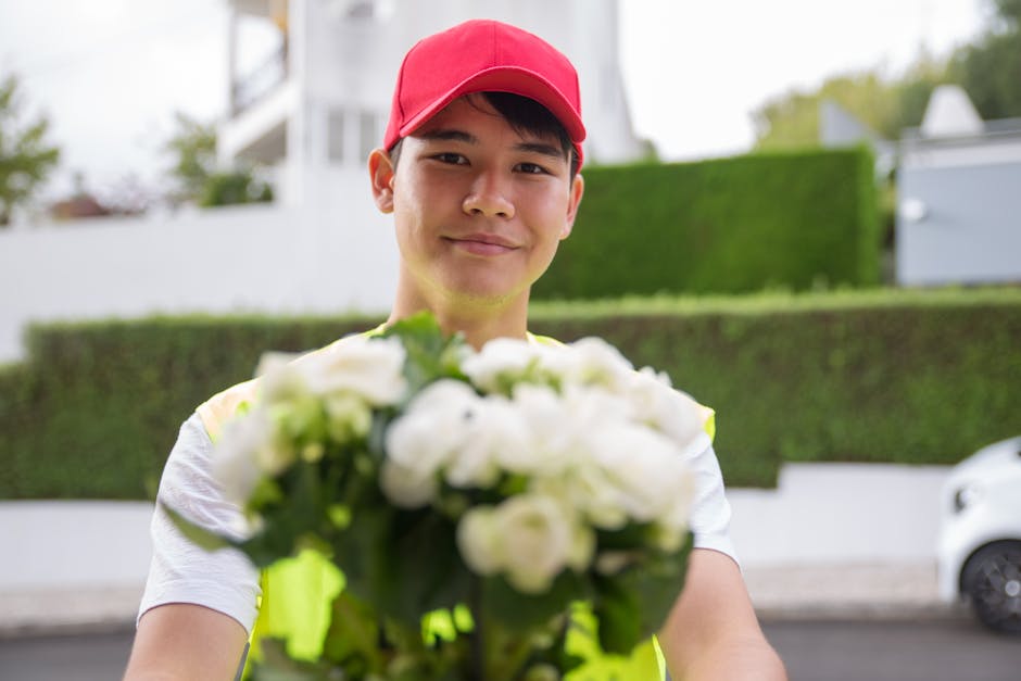 A young male flower delivery courier wearing a red cap, holding a bouquet of fresh white roses and green foliage, with a cheerful expression. He is outdoors in a residential area, standing in front of