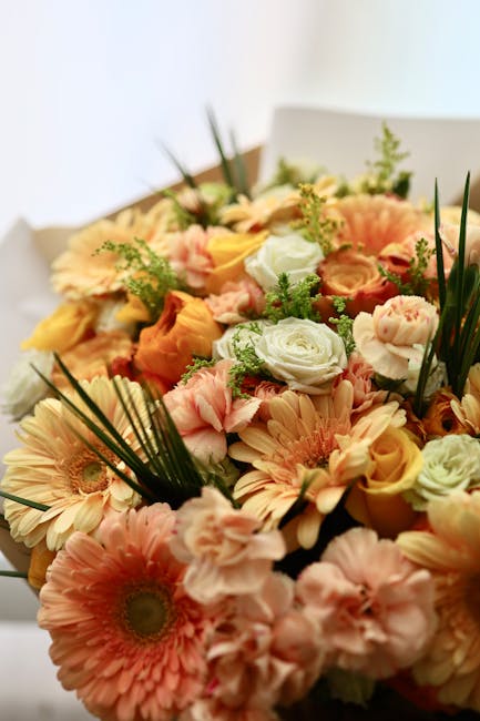 A vibrant bouquet of fresh flowers featuring peach and cream gerbera daisies, pale pink carnations, soft yellow roses, and white roses with green foliage and long, slender leaves. The arrangement is w